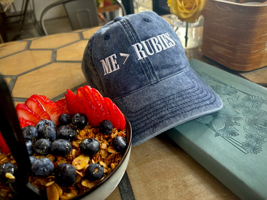 Blue cap with 'ME > RUBIES' text next to a bowl of fruit and granola on a table.