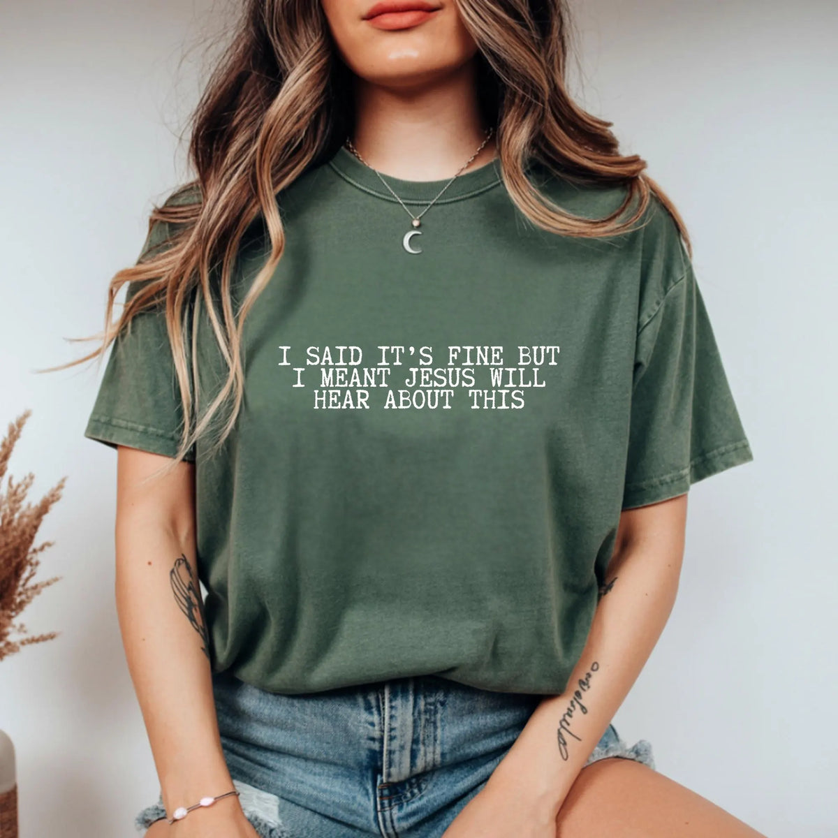 Person wearing a green t-shirt with text "I said it's fine but I meant Jesus will hear about this" sitting against a neutral background