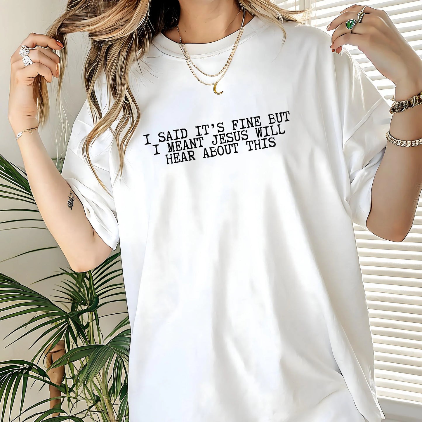 Person wearing a white t-shirt with humorous text design "I said it's fine but I meant Jesus will hear about this" standing indoors with plants in the background.
