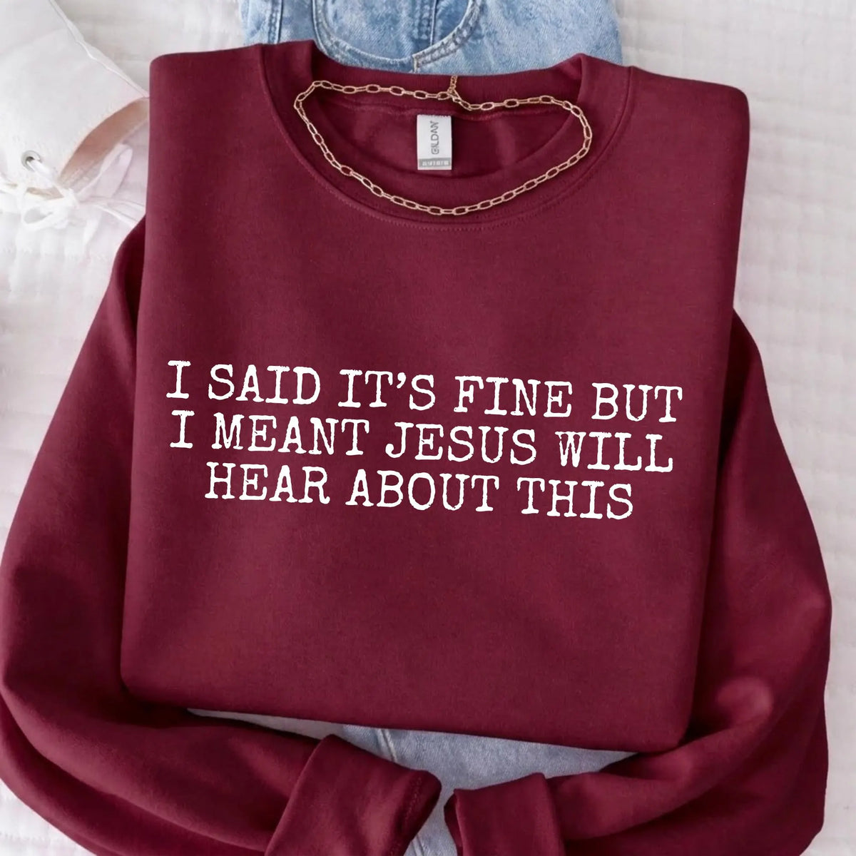 Maroon sweatshirt with humorous text "I said it's fine but I meant Jesus will hear about this" on a light background