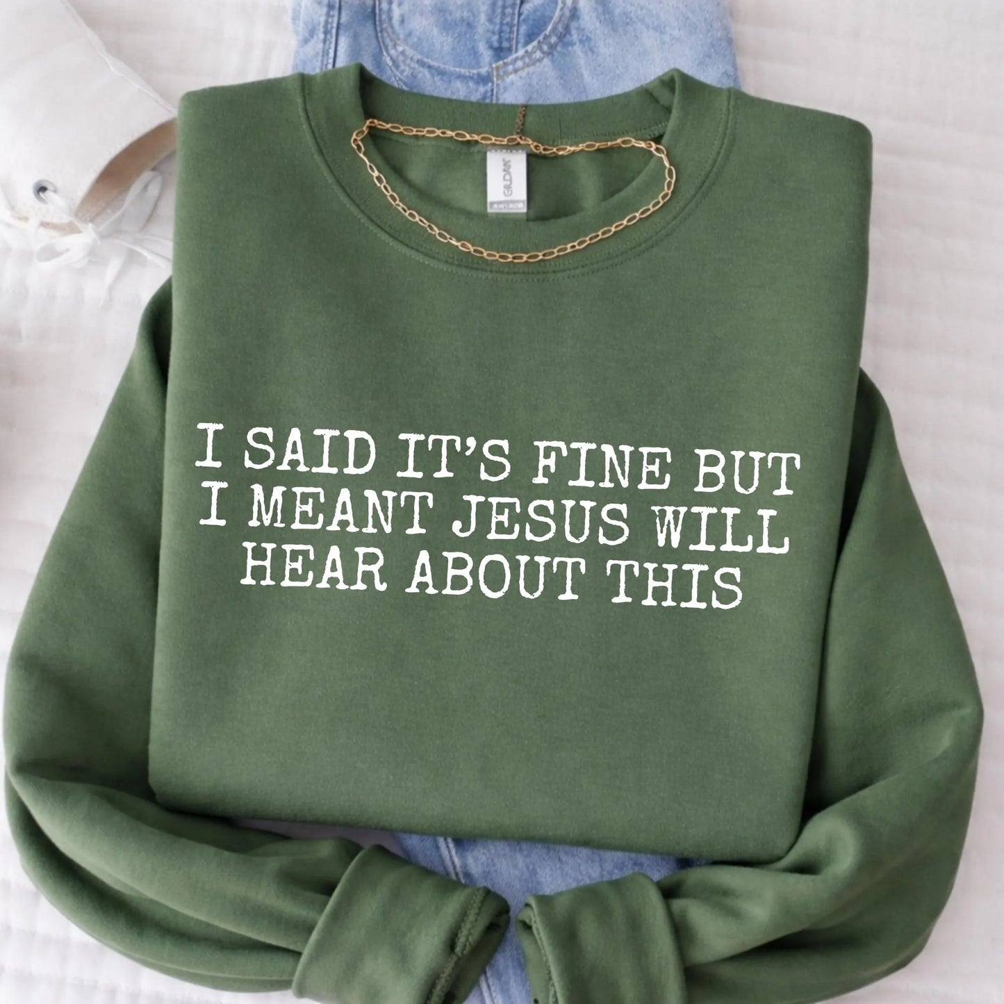 Green sweatshirt with text 'I said it's fine but I meant Jesus will hear about this' on a light background
