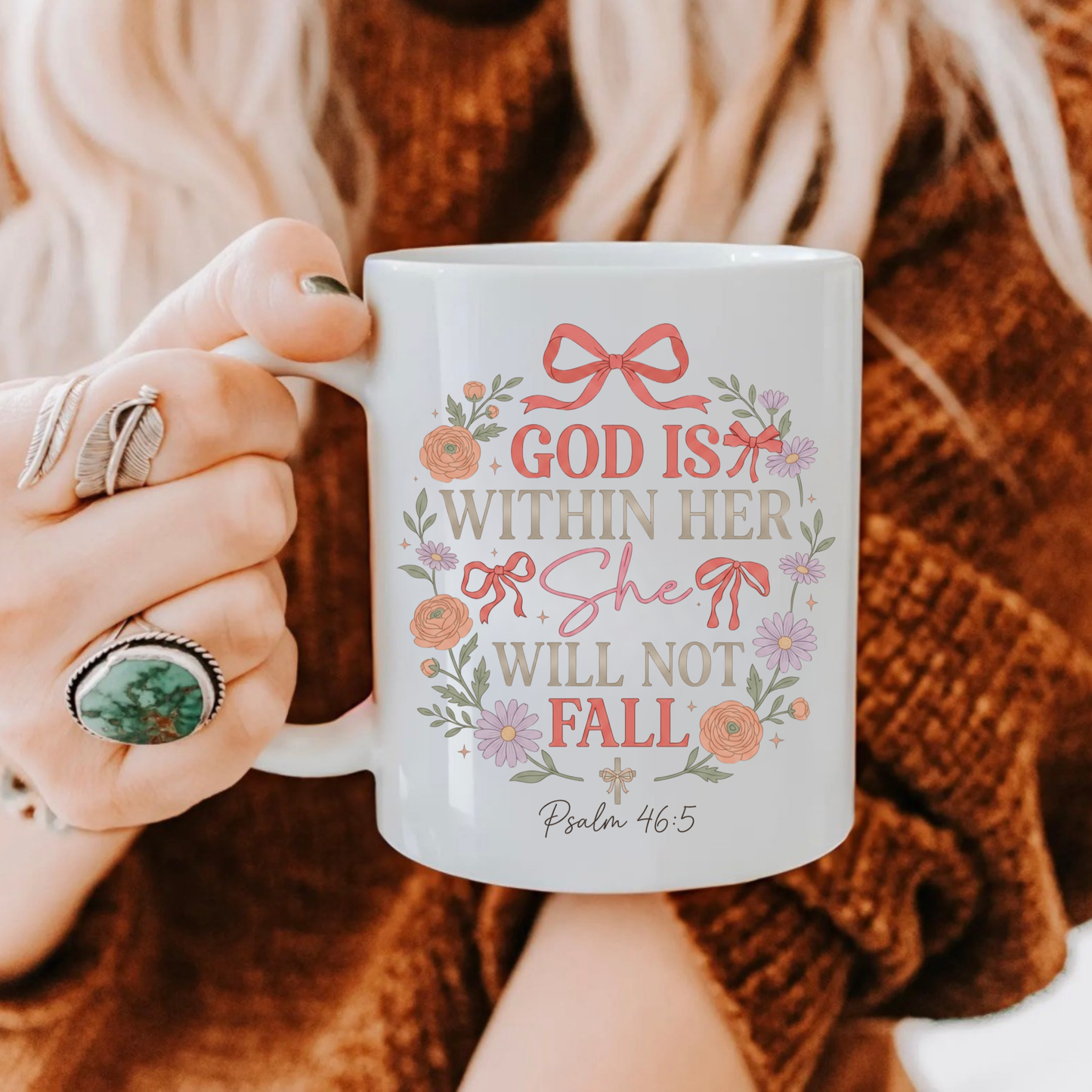 Person holding a mug with decorative text 'God is within her, she will not fall' and design, wearing a brown sweater.