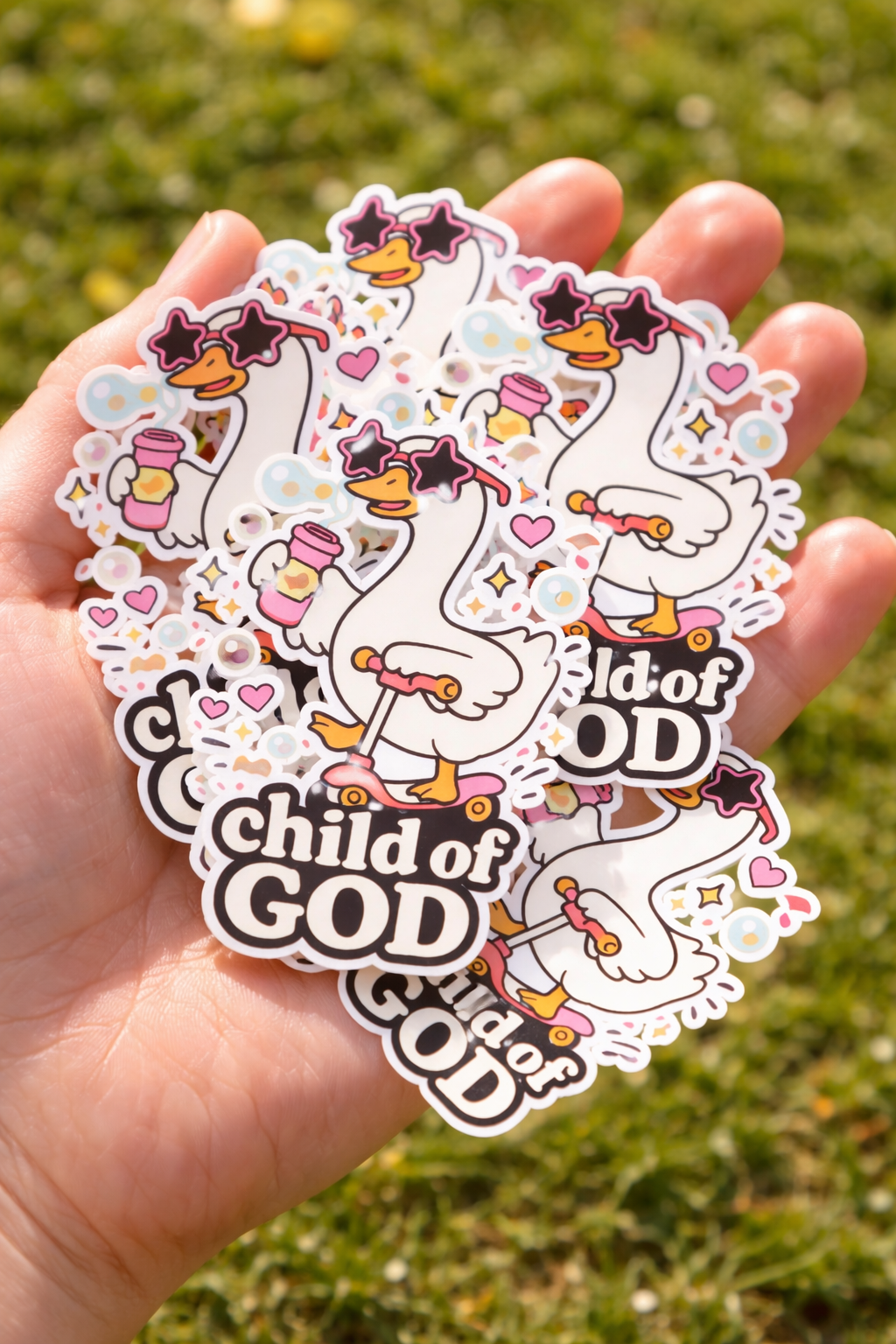 Hand holding a stack of 'Child of God' themed stickers with cartoon goose on a grassy background.