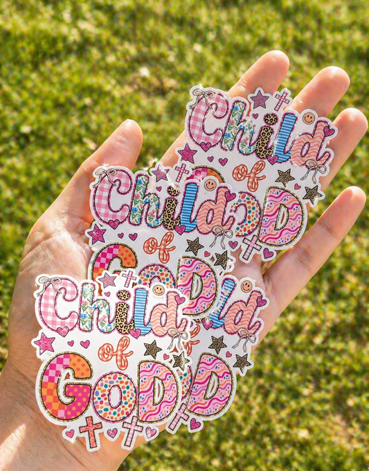 Hand holding three cute Child of God Christian stickers with colorful patchwork letters, crosses, hearts, and stars in a bright outdoor setting.
