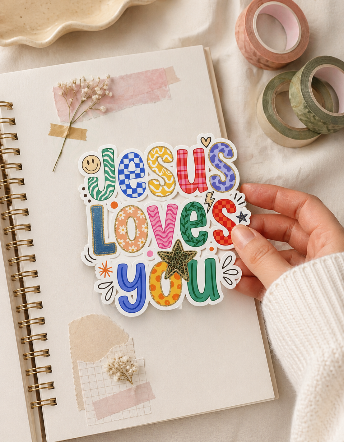 Hand holding colorful Jesus loves you Christian sticker over aesthetic scrapbook journal with soft neutral background, floral details, and washi tape, cute faith sticker mockup for laptop, water bottle, journal, and Bible journaling.