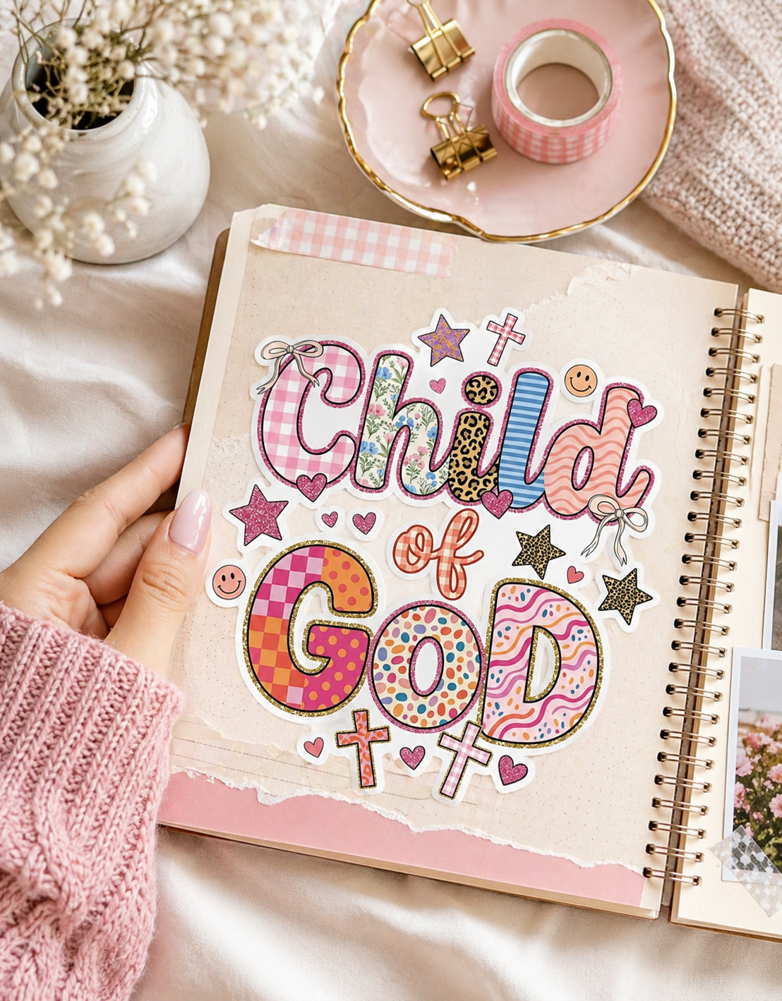 Cute Child of God sticker with colorful patchwork lettering placed on an open scrapbook journal, surrounded by pink stationery, washi tape, gold binder clips, and soft floral decor in a cozy aesthetic flat lay.