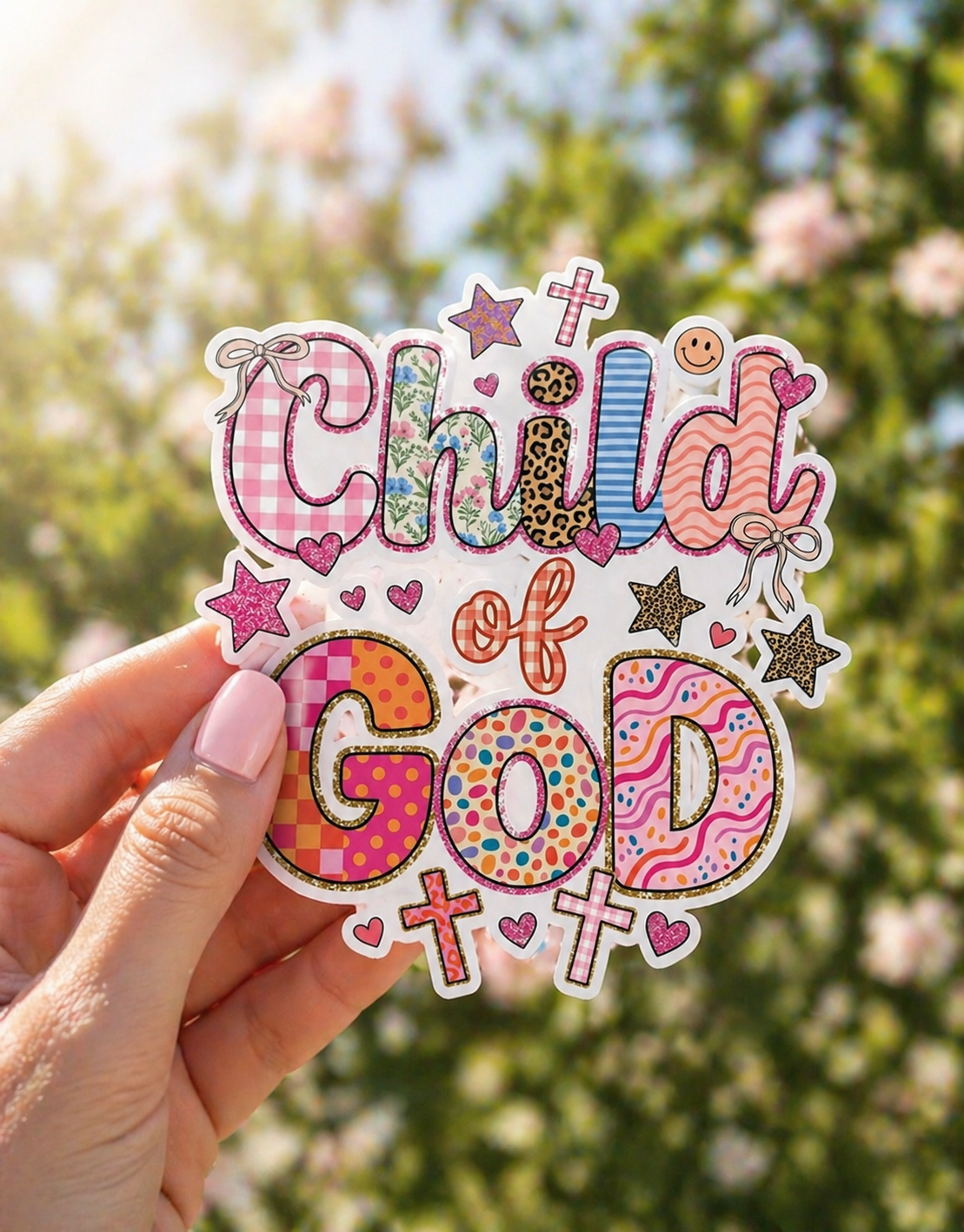 Child of God Christian sticker held outdoors with colorful patchwork design, crosses, hearts, and a soft floral spring background.