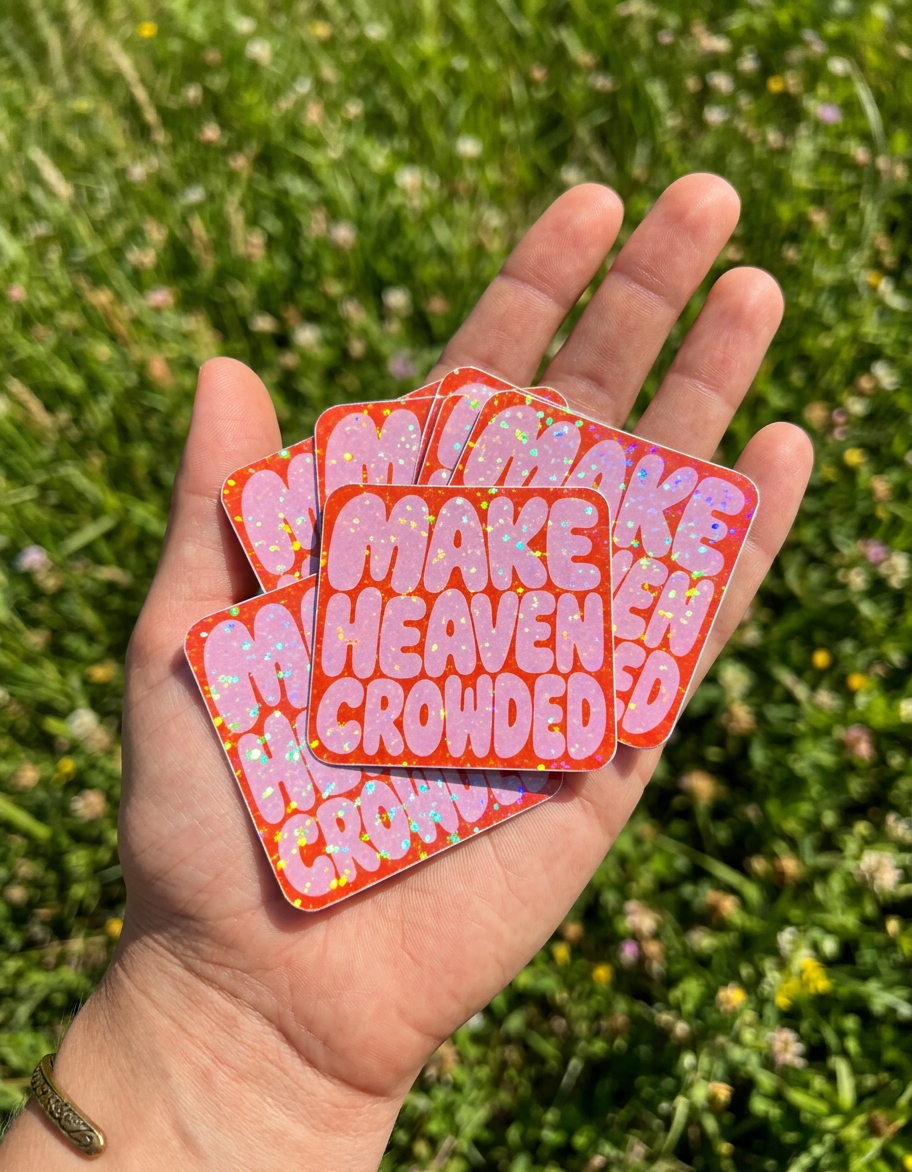 Hand holding three colorful stickers with 'MAKE HEAVEN CROWDED' text against a grassy background