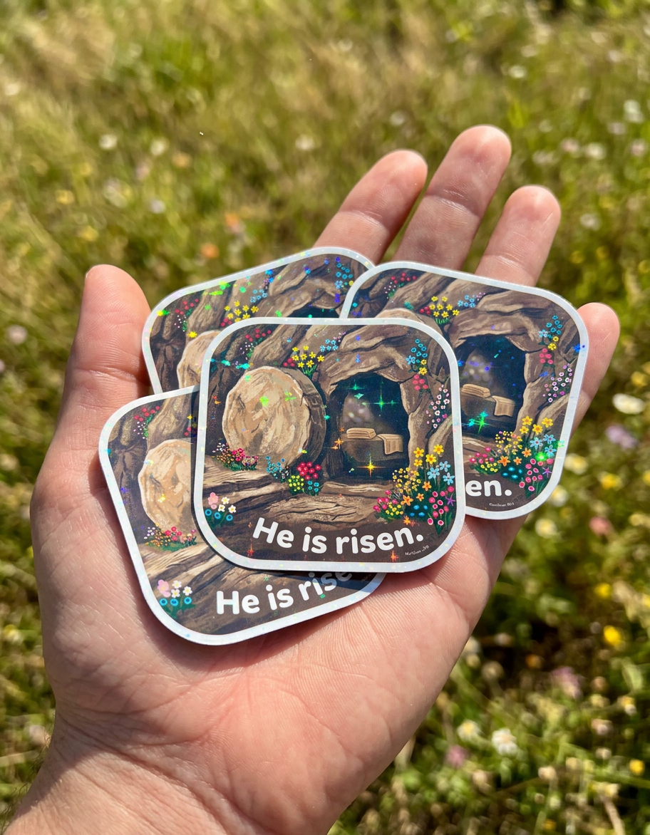 An open hand holds several square holographic stickers with rounded corners, each featuring an illustrated empty tomb surrounded by colorful flowers. The stone is rolled away from the cave entrance, folded cloths rest inside, and white text at the bottom reads, “He is risen.” Sunlight catches the stickers, creating sparkling reflections against a blurred grassy background.