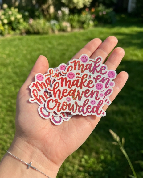 An open palm holds a cluster of holographic, glittery stickers that read "make heaven crowded." in a red script with pink smiley faces. The person is wearing a delicate silver cross bracelet, and the background shows a bright, sunlit green lawn and garden.