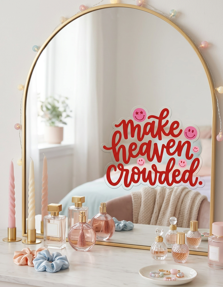 A die-cut sticker with the phrase "make heaven crowded." in red script is attached to the center of an arched gold vanity mirror. The mirror is decorated with a string of colorful fairy lights and sits on a marble surface alongside perfume bottles, twisted pastel candles, and blue and peach scrunchies.