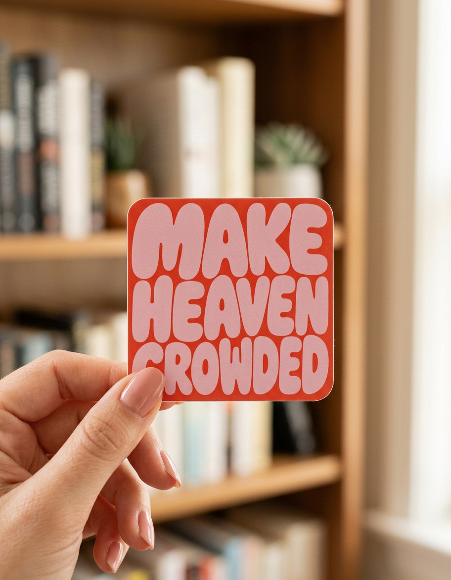 Hand holding a red sticker with 'MAKE HEAVEN CROWDED' text in front of a bookshelf.