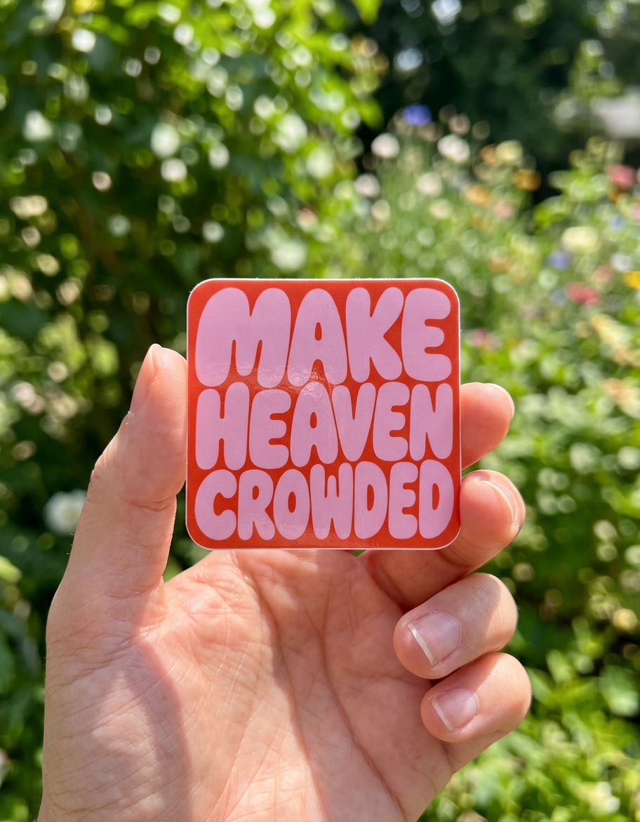 Hand holding a pink sticker with 'MAKE HEAVEN CROWDED' text against a blurred green background