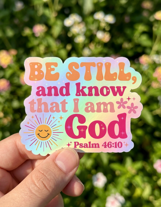 Holographic sticker with 'Be still and know that I am God' text held in front of green foliage.