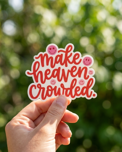 A person's hand holds a die-cut sticker outdoors against a blurred green leafy background with soft bokeh. The white sticker features the phrase "make heaven crowded." in a red cursive font, accented by several small pink smiley faces with star and dot eyes.