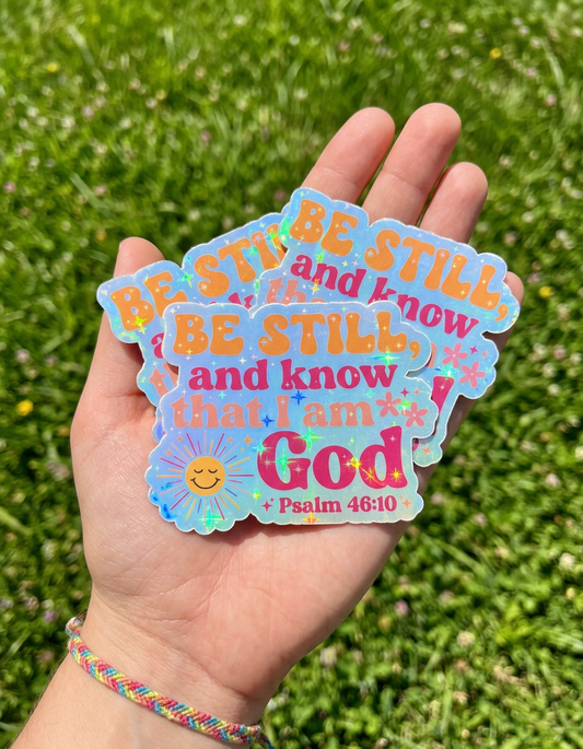 Hand holding colorful 'Be Still and Know That I Am God' stickers against a grass background