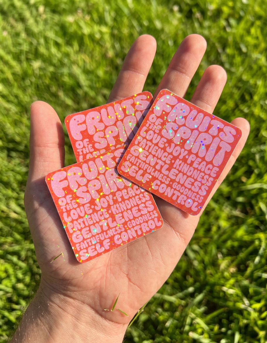 An open palm holds three square "Fruits of the Spirit" stickers with a shimmering, holographic glitter finish. The vibrant orange and pink designs are shown outdoors against a background of bright green grass in the sunlight.