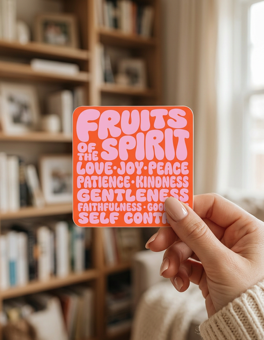 A person’s hand holds a square sticker with rounded corners against a blurred background of a wooden bookshelf. The sticker has a vibrant orange background with bubbly, pink text that reads "FRUITS OF THE SPIRIT" followed by a list of virtues: "LOVE, JOY, PEACE, PATIENCE, KINDNESS, GENTLENESS, FAITHFULNESS, GOODNESS, SELF CONTROL.