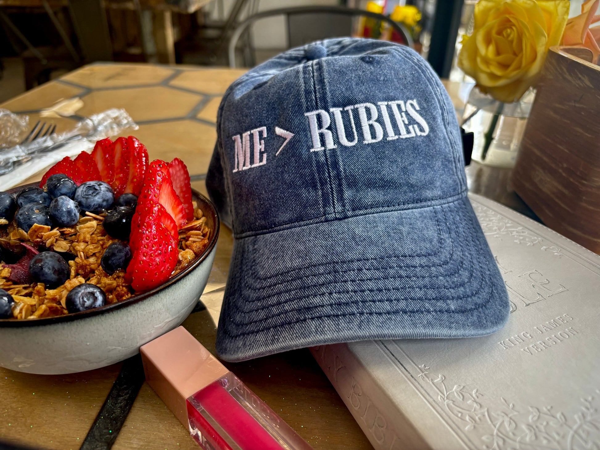 Blue cap with 'ME RUBIES' text on a table with a fruit bowl and pen.