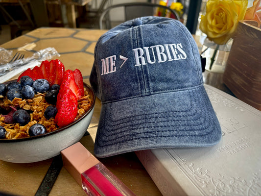 Blue cap with 'ME RUBIES' text on a table with a fruit bowl and pen.