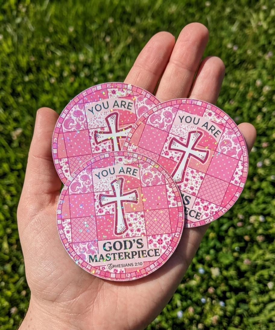 Pink stickers "You Are God's Masterpiece" with cross design held in a hand against a grass background