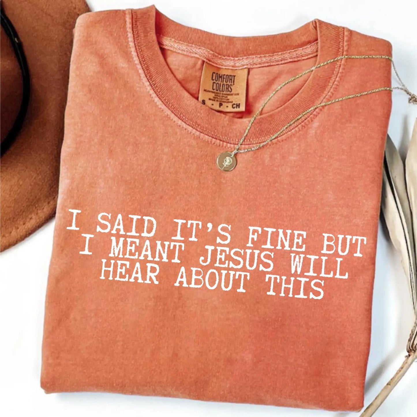 Folded orange t-shirt with humorous text "I said it's fine but I meant Jesus will hear about this" on a white surface