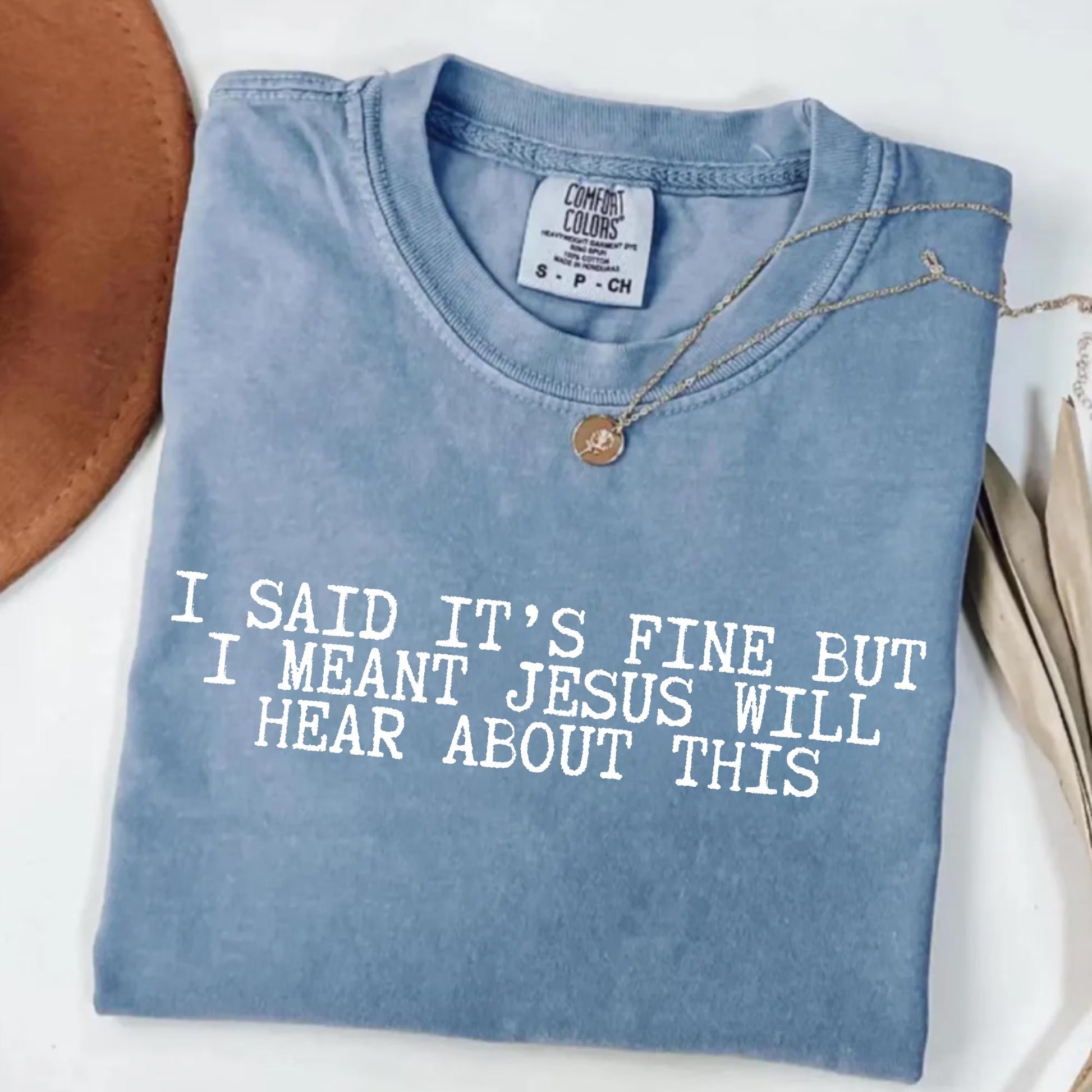 Folded blue t-shirt with humorous text "I said it's fine but I meant Jesus will hear about this" on a white surface