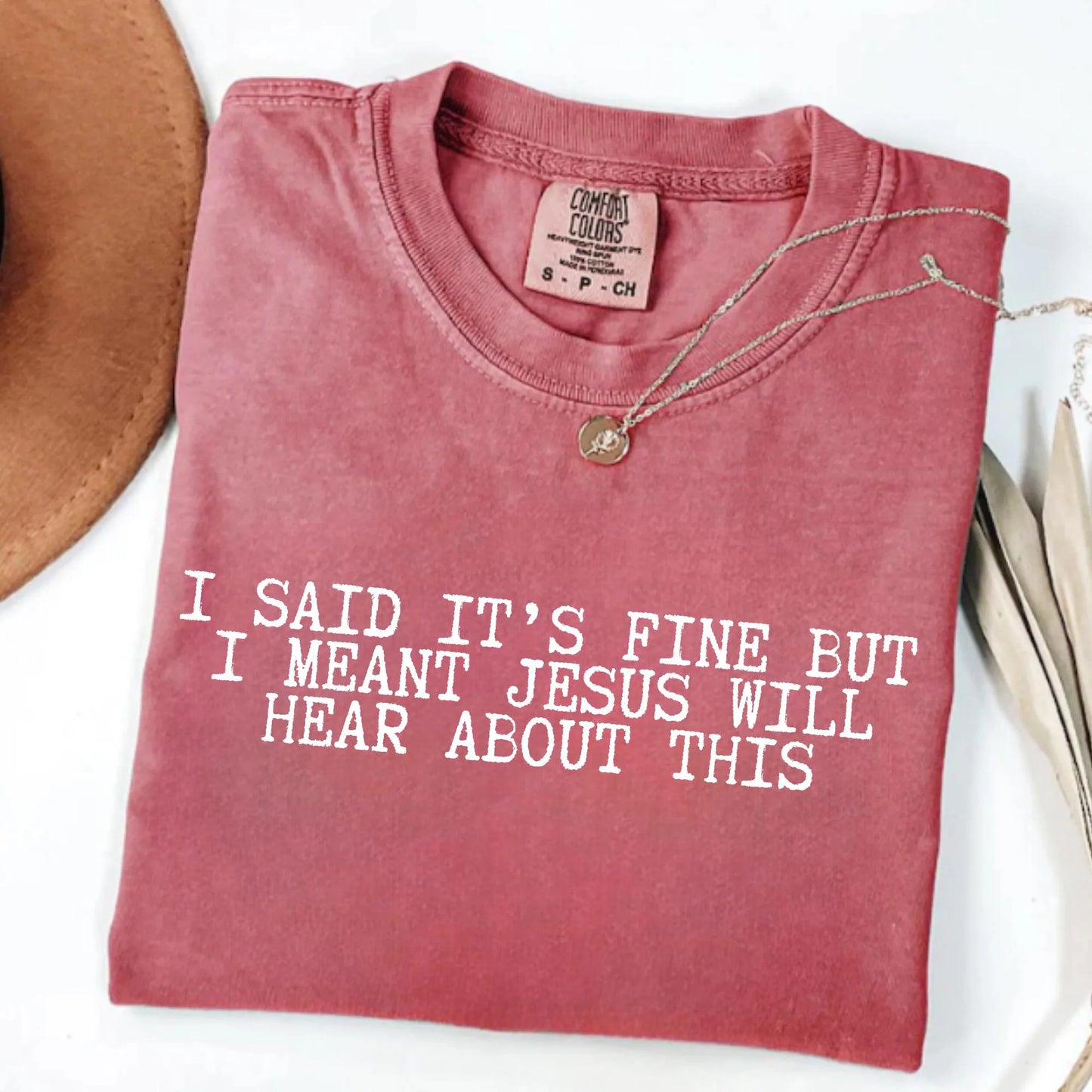 Folded red t-shirt with humorous text "I said it's fine but I meant Jesus will hear about this" on a white surface