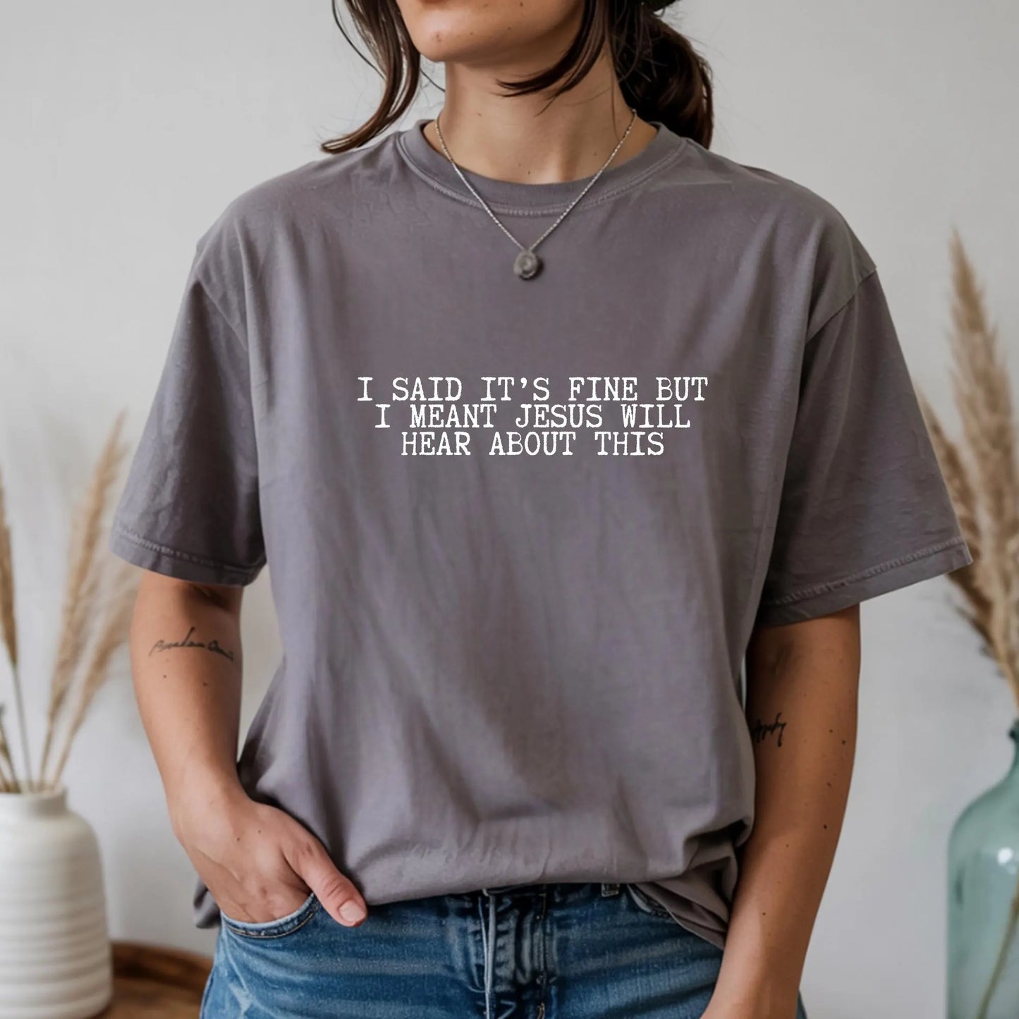 Person wearing a gray t-shirt with a humorous text "I said it's fine but I meant Jesus will hear about this" design.