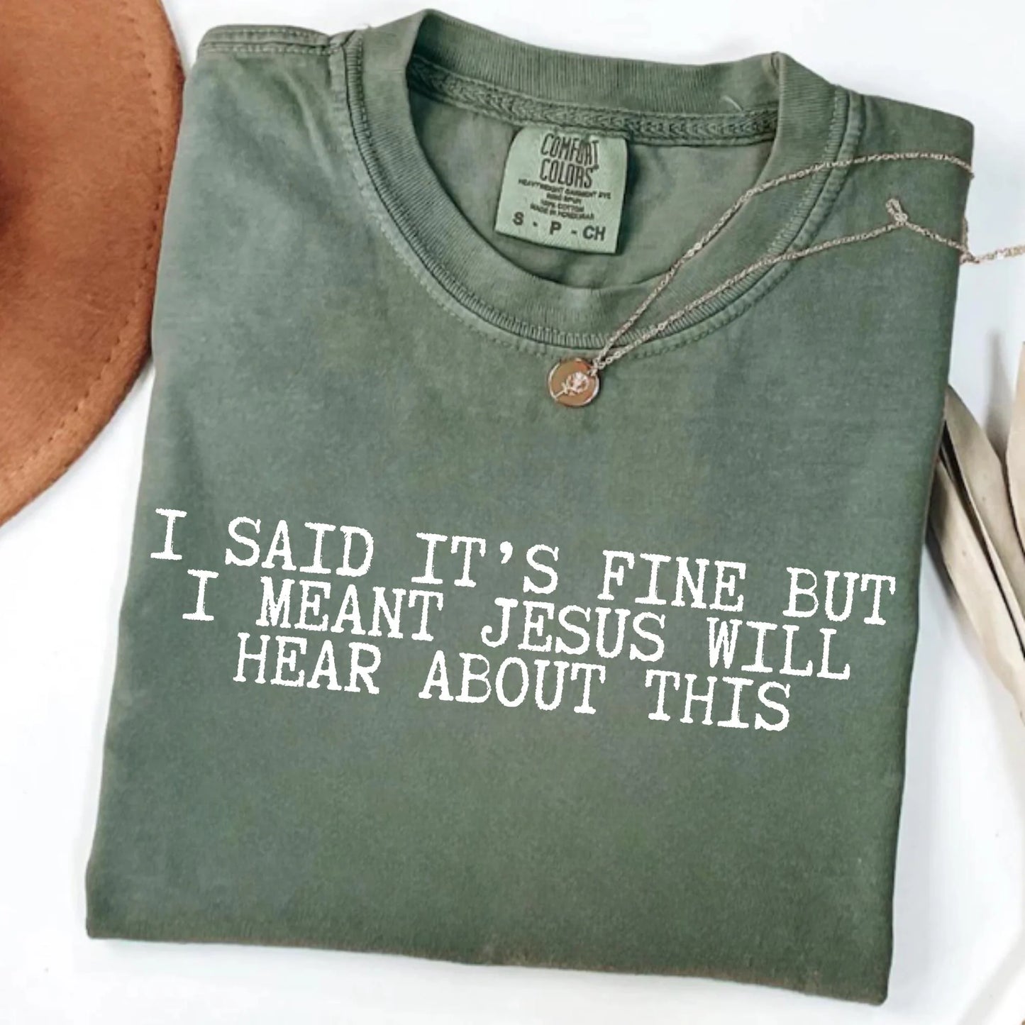 Folded green t-shirt with humorous text "I said it's fine but I meant Jesus will hear about this" on a white background