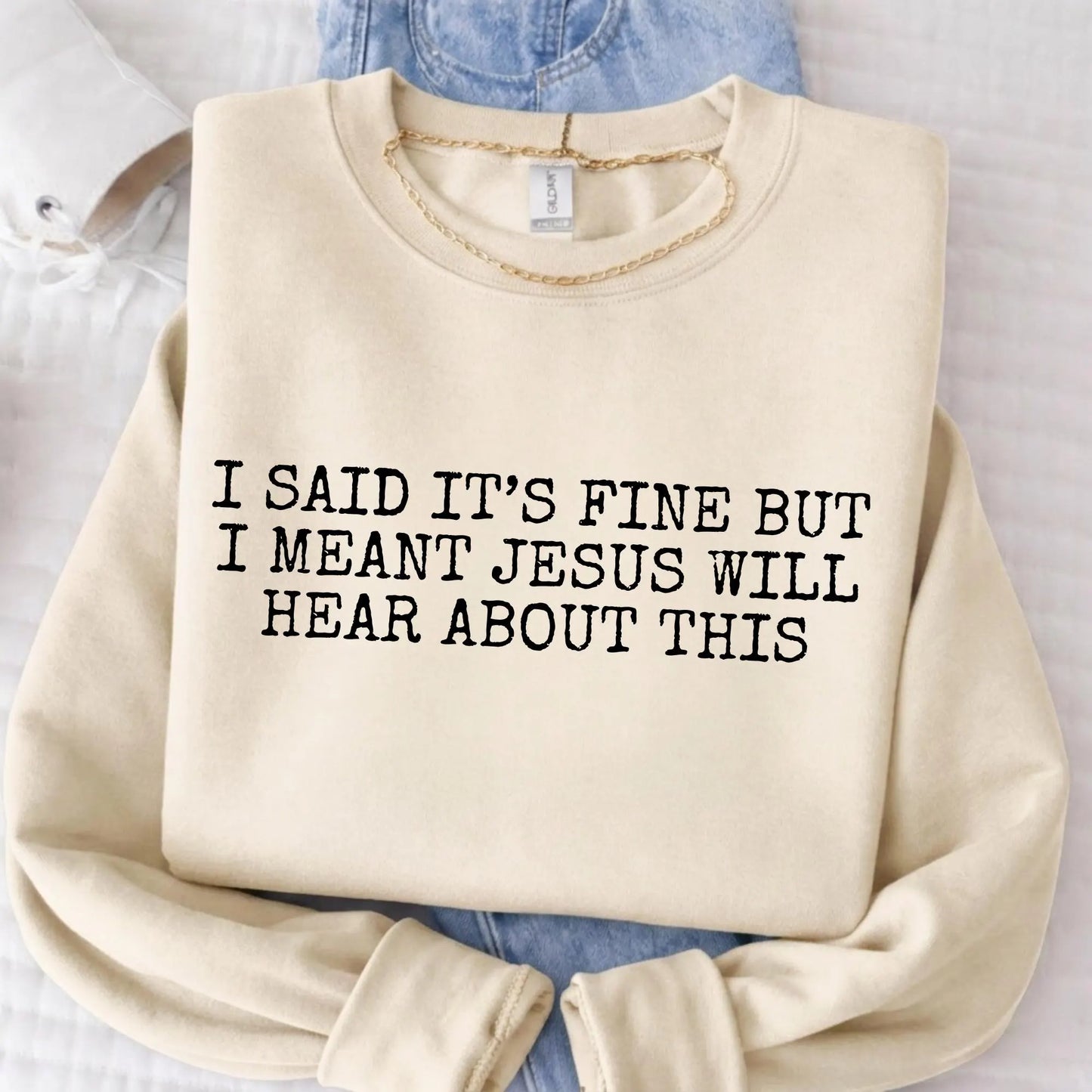 Beige sweatshirt with text 'I said it's fine but I meant Jesus will hear about this' on a light background