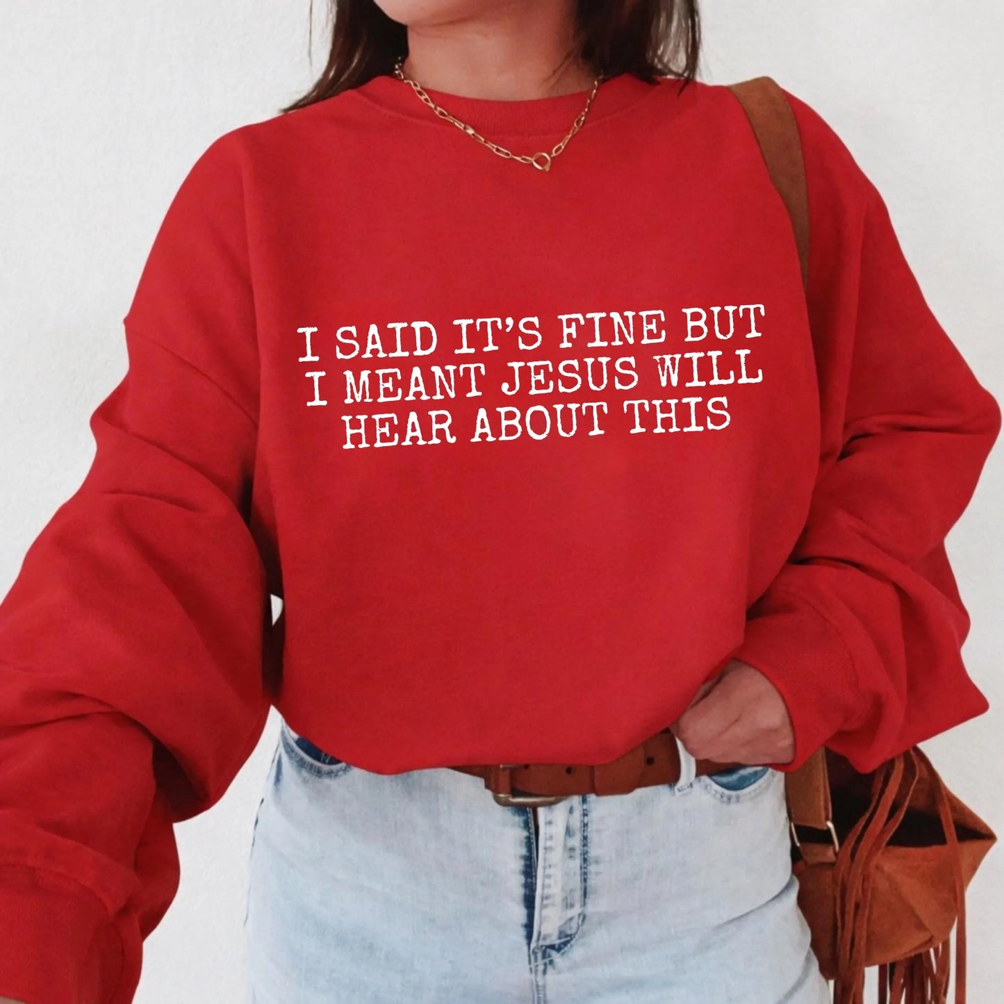 Person wearing a red sweatshirt with humorous text "I said it's fine but I meant Jesus will hear about this" design on a plain background
