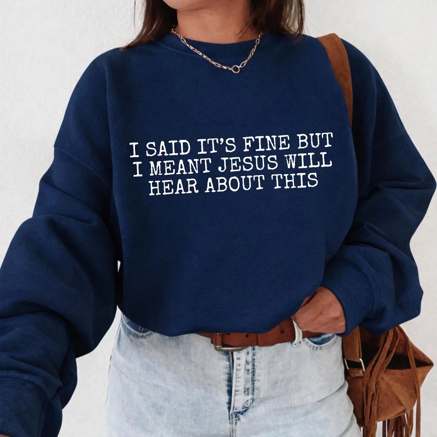 Person wearing a navy blue sweatshirt with humorous text "I said it's fine but I meant Jesus will hear about this", standing against a plain background.
