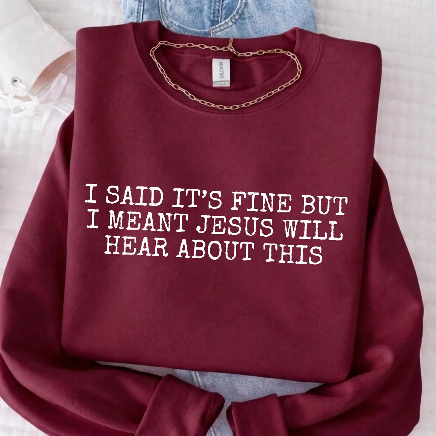 Maroon sweatshirt with humorous text "I said it's fine but I meant Jesus will hear about this" on a light background