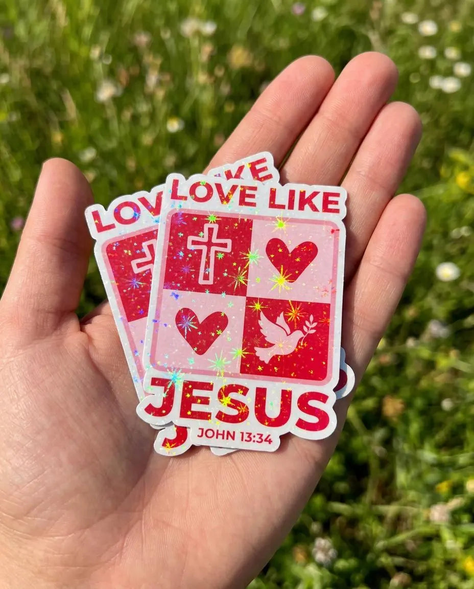 Hand holding a christian sticker "Love like Jesus" "John 13:34" against a green grass background