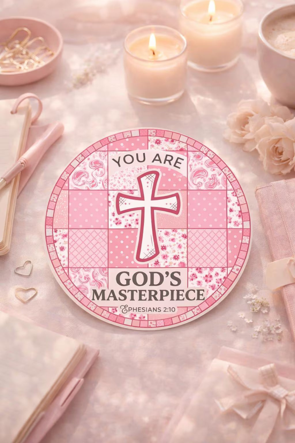 Decorative sticker with a cross design and text 'You are God's Masterpiece' on a pink surface with candles and flowers.