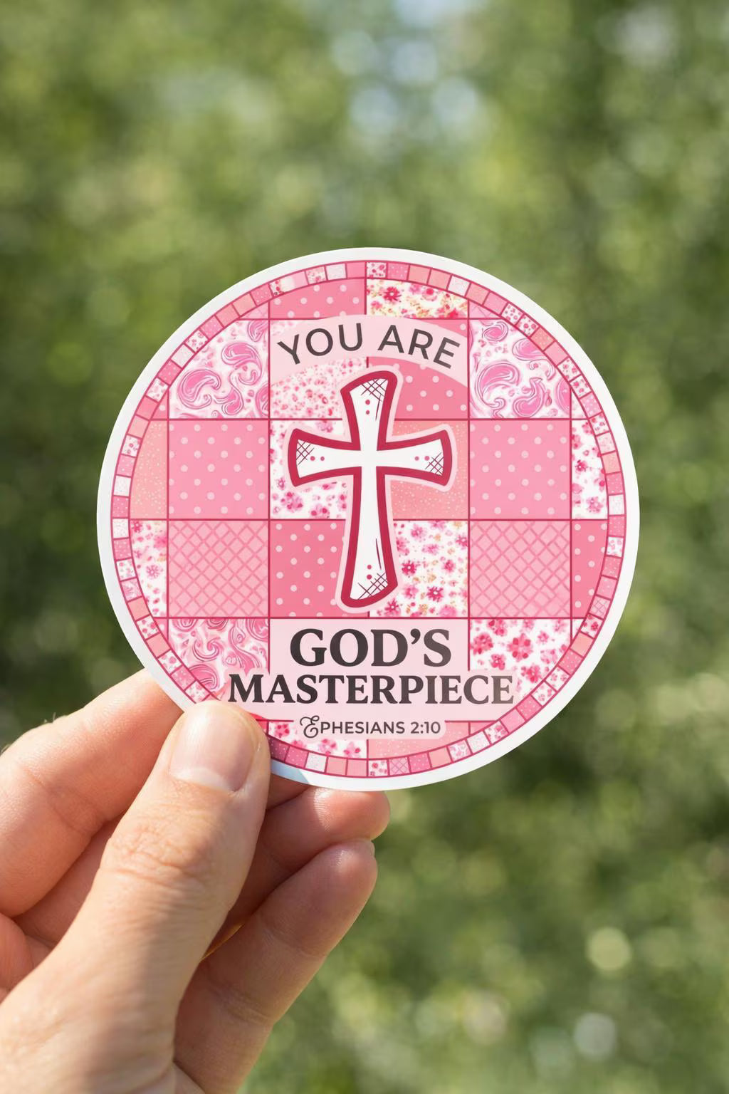 Pink sticker "You Are God's Masterpiece" with cross design held by a hand against a blurred green background