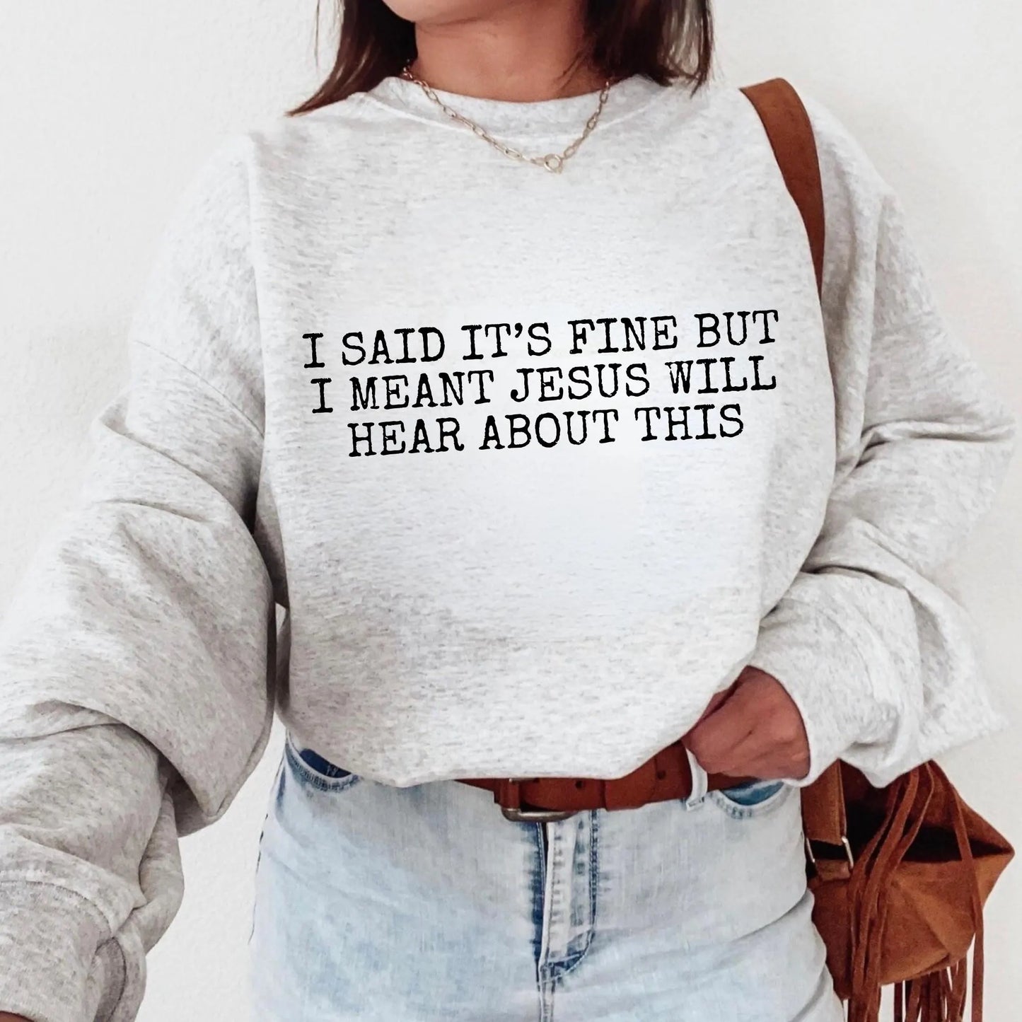 Person wearing a gray sweatshirt with text "I said it's fine but I meant Jesus will hear about this" standing against a white background