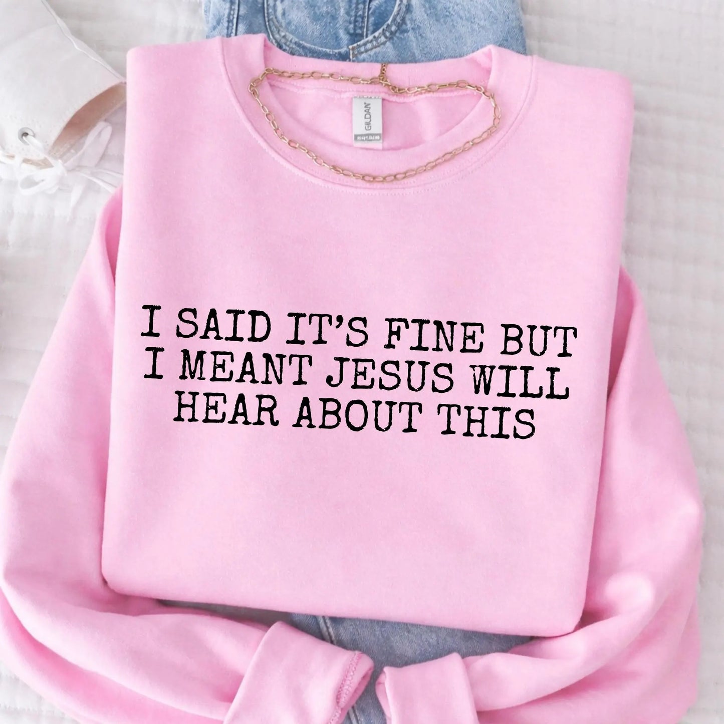 Pink sweatshirt with humorous text "I said it's fine but I meant Jesus will hear about this" on a light background
