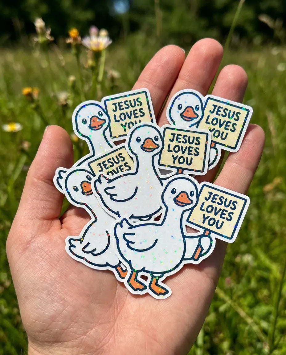 Hand holding stickers with geese and 'Jesus Loves You' sign on hand against a grassy background